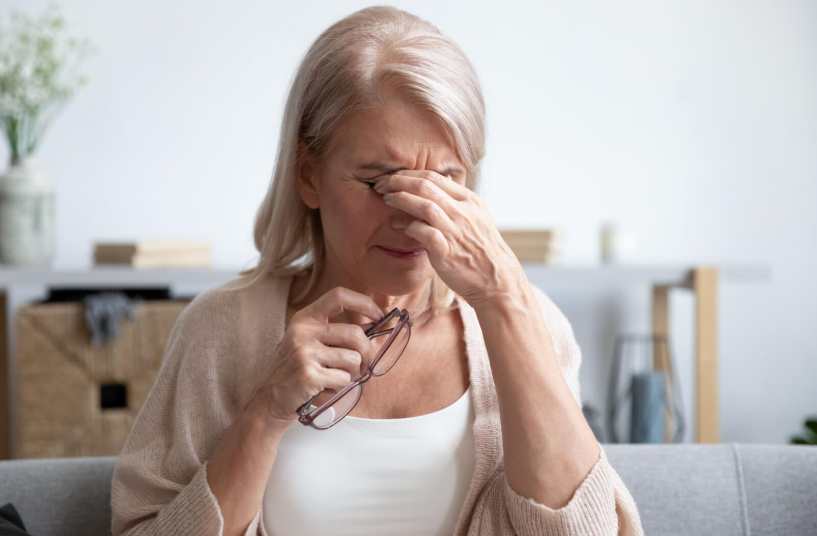 A senior woman sitting on a couch while holding her glasses in her right hand as she rubs her eyes.
