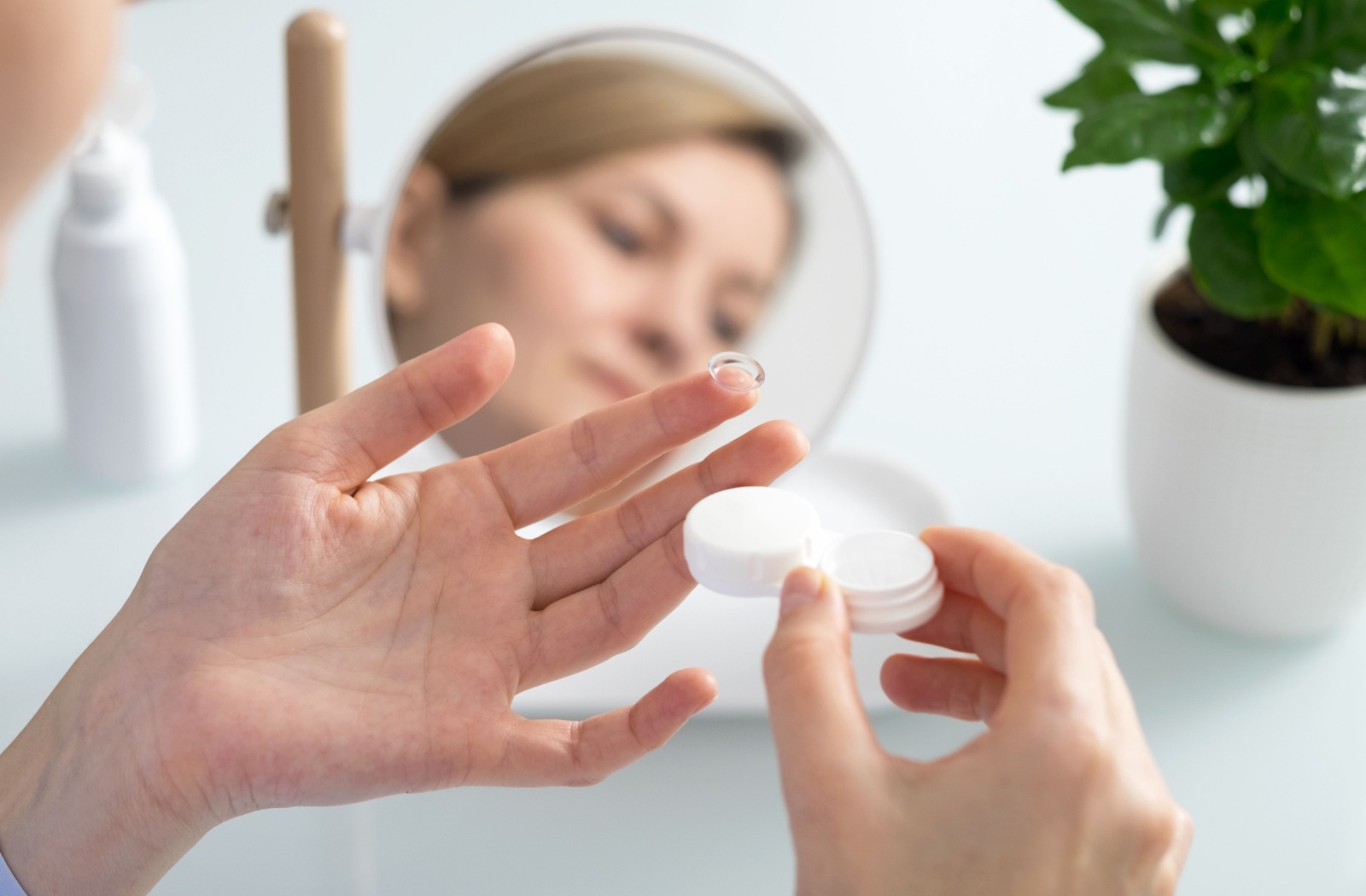 A woman holds a contact lens on her left index finger, and a contact lens case with solution in her right hand.