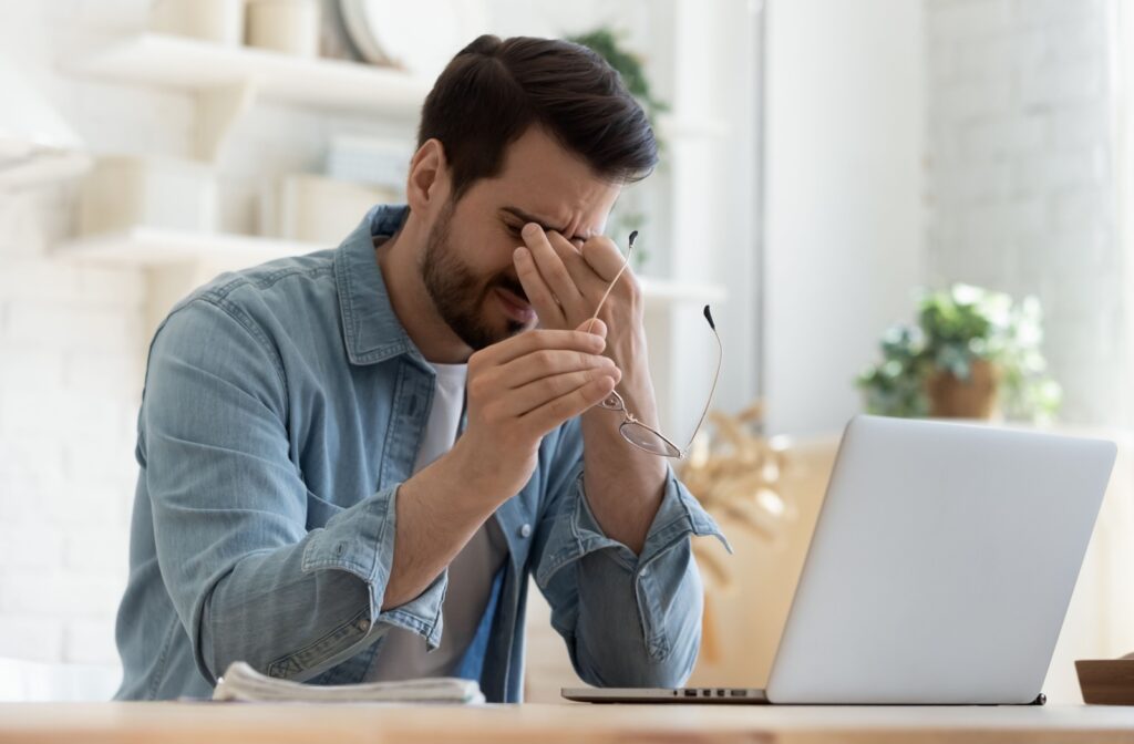 A young man taking off his glasses while working to rub his eyes due to dry eye discomfort.