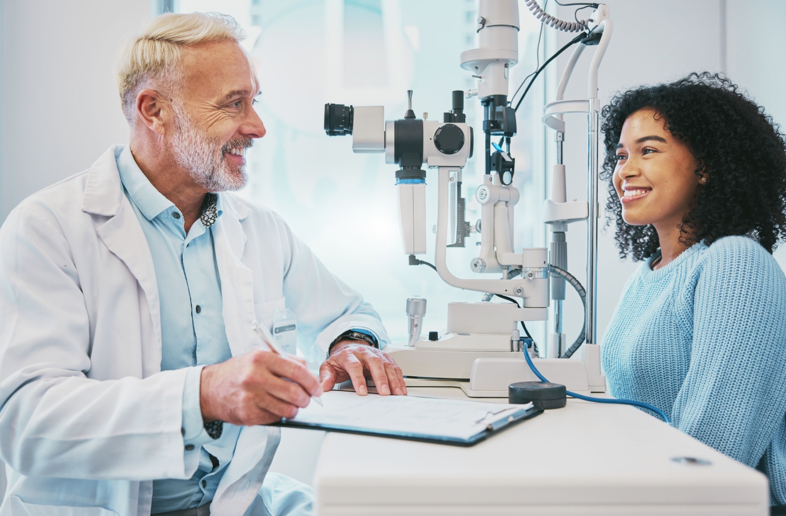 An optometrist and his patient smiling across the table while discussing the results of her eye exam.
