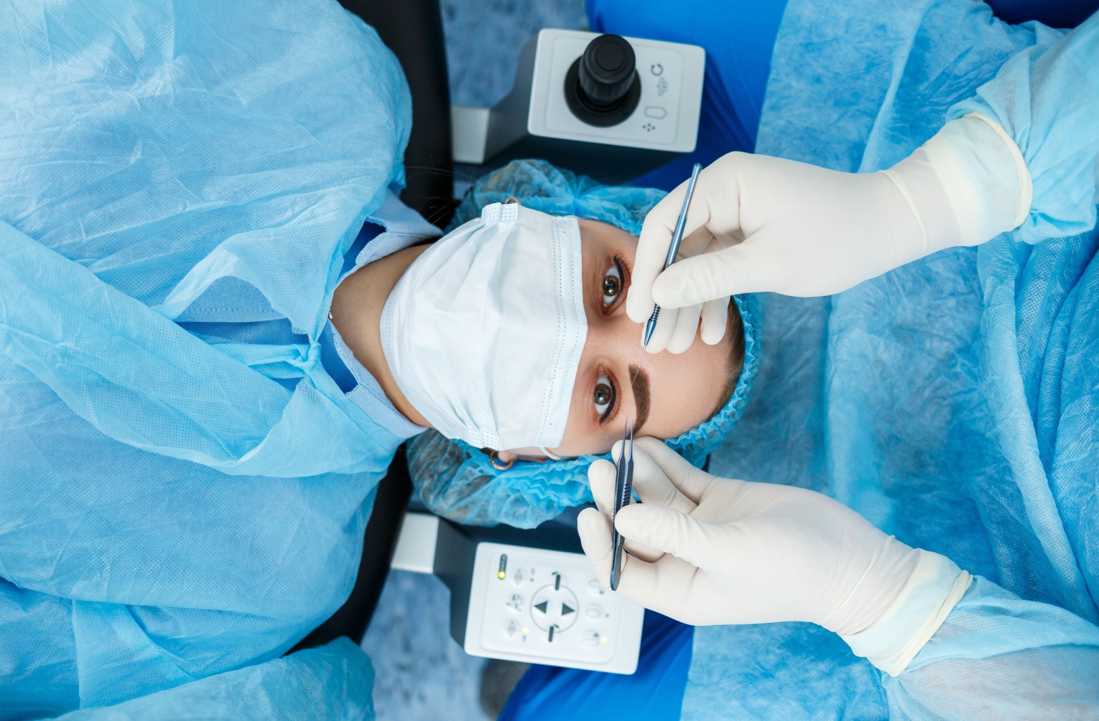 A laser eye surgery patient looks up with wide eyes at a technician's hands holding medical tools