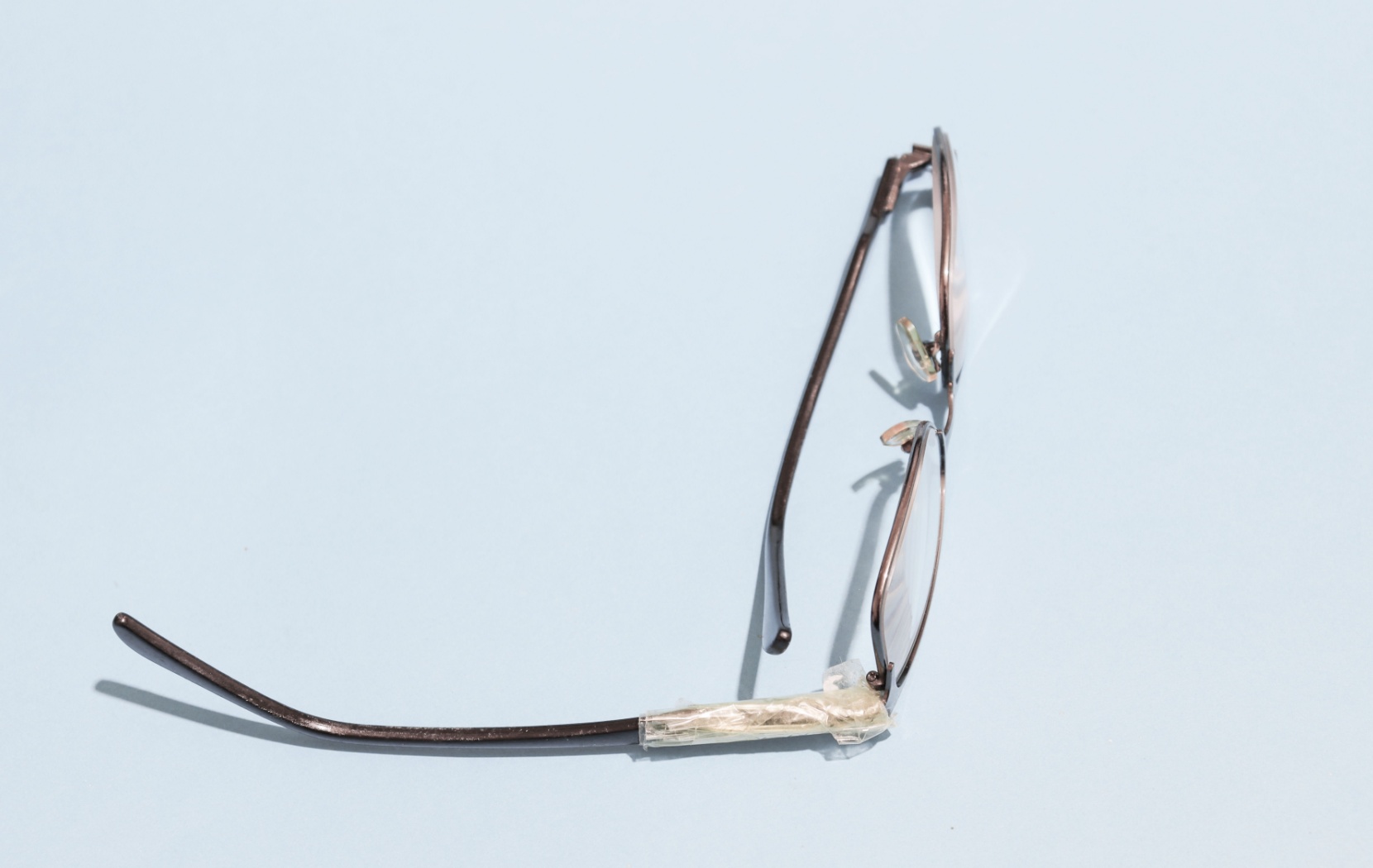 A pair of eyeglasses with a broken, taped arm sits on a pale blue background with the non-broken arm folded in.