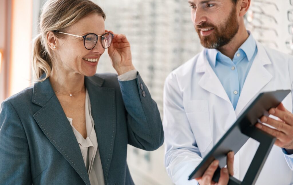 An optometrist holds up a mirror to show their patient a pair of glasses during a an eyeglasses fitting appointment.