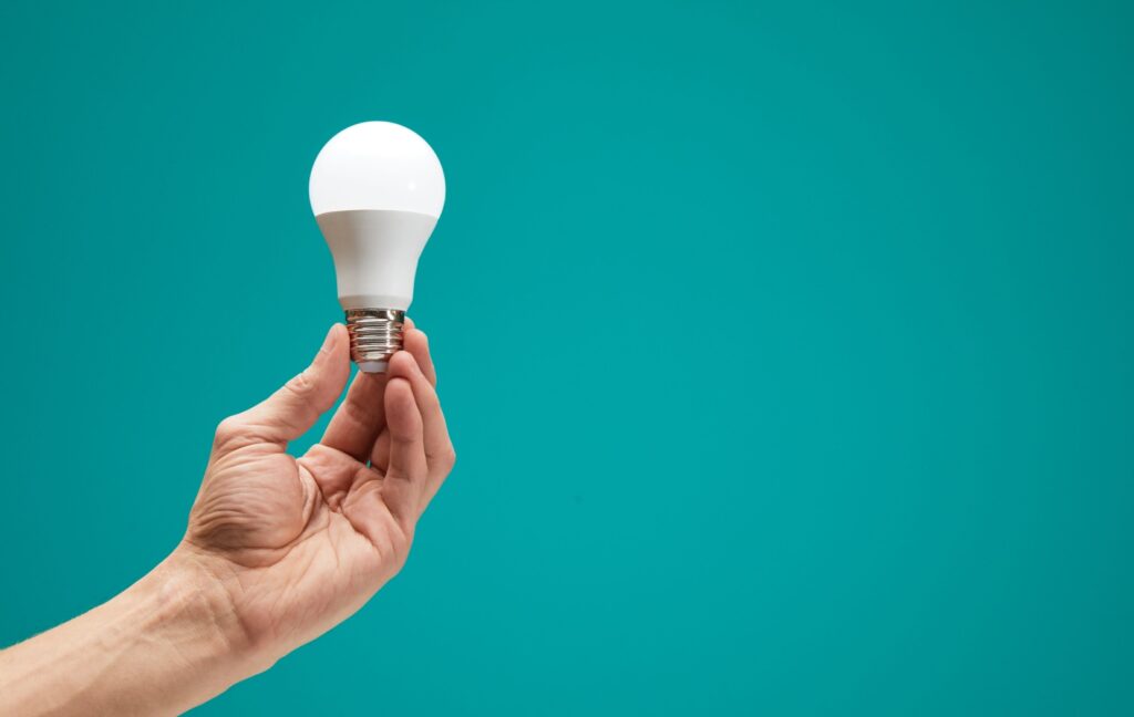 A person holds up a white LED light bulb in front of a teal colored background.
