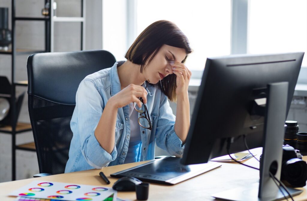 Woman sitting at her desktop computer holding her glasses in her hand while rubbing her eyes.