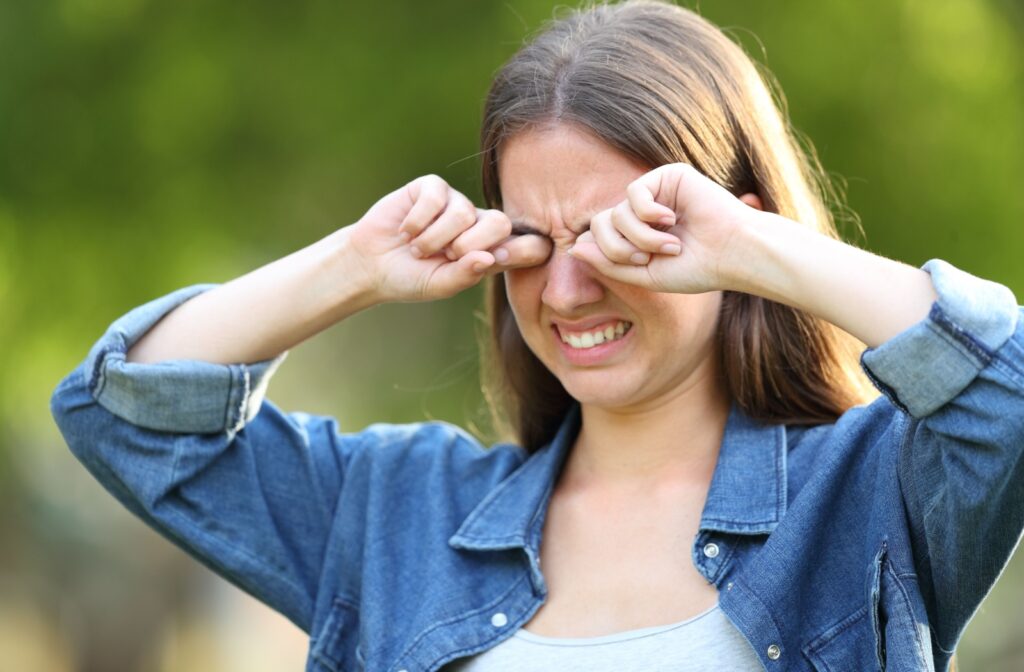 Young woman outdoors rubbing her eyes with both hands, showing discomfort and irritation commonly caused by allergies or dry eyes.