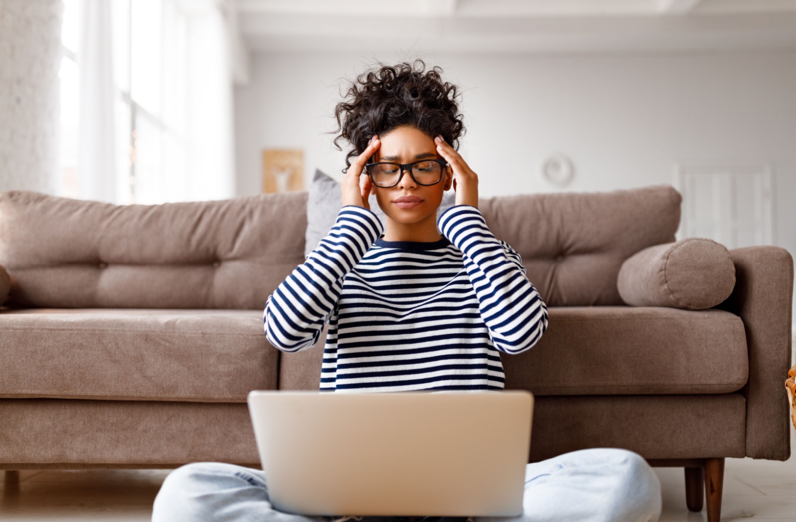 Person sitting on the floor using a laptop, holding their head due to a headache caused by symptoms of dry eye.