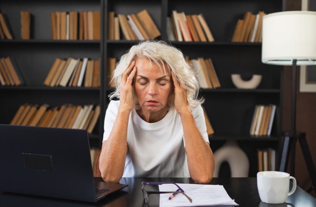 A person sitting at a desk, holding their head because they are experiencing a headache caused by the symptoms of dry eye disease.
