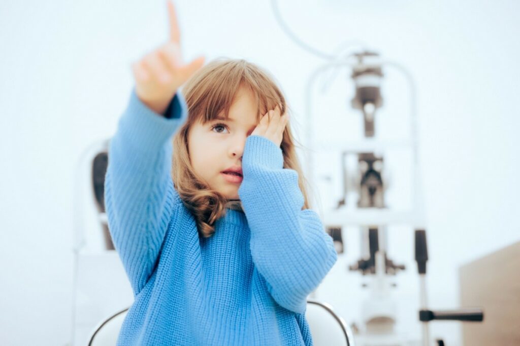 A young girl in a blue sweater covering one eye and pointing her finger while getting a vision test at an ophthalmologist's office.
