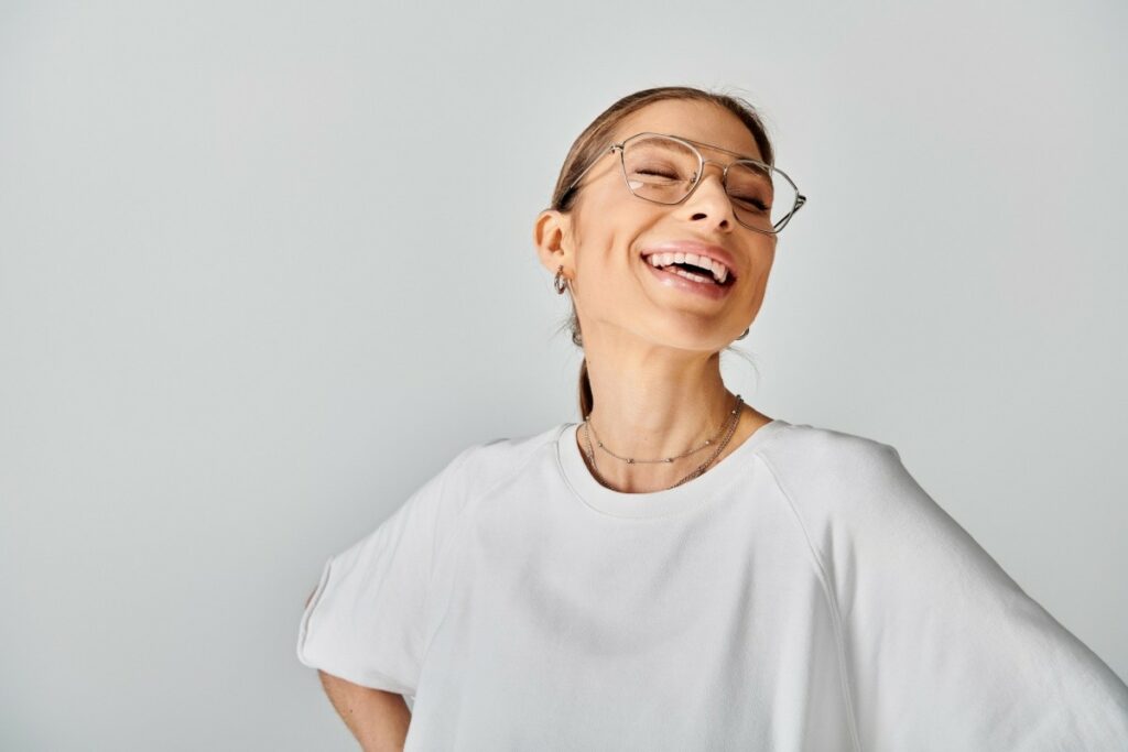 A joyful young woman with brown hair tied back, wearing stylish geometric glasses and a white t-shirt, laughing heartily with her eyes closed against a neutral gray background.