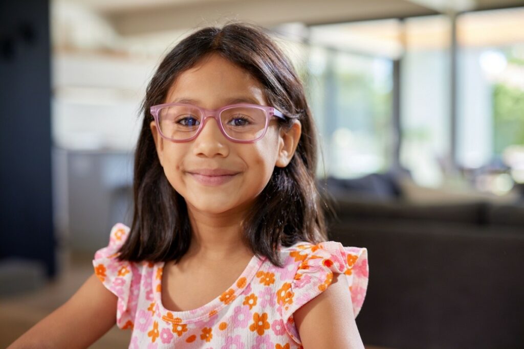 A young girl with dark hair and pink-framed glasses smiles cheerfully while wearing a pink floral top in a sunny indoor room.