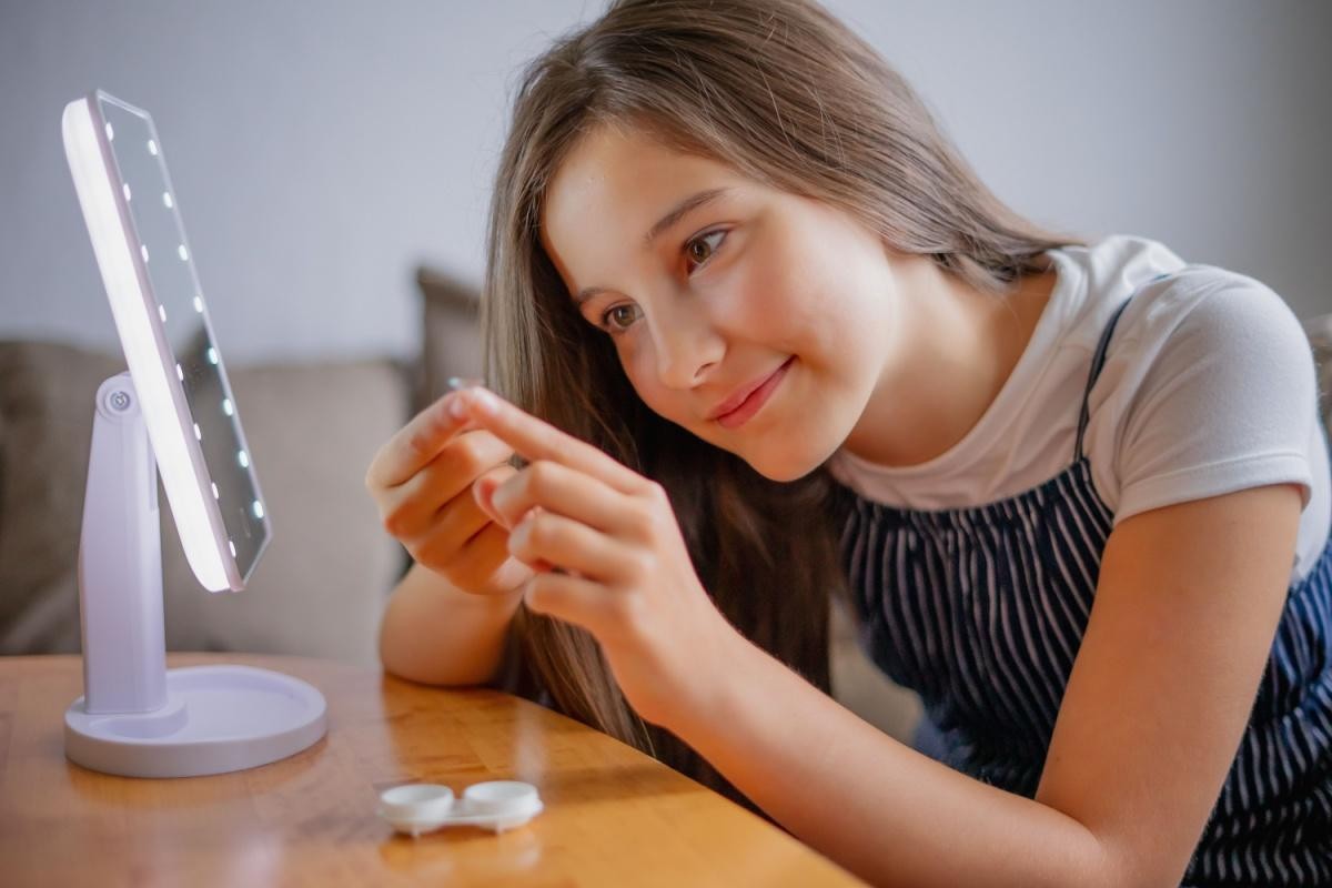 A young girl with long brown hair carefully holds a contact lens on her finger as she prepares to insert it, using a small lighted vanity mirror on a table to assist her with the process at Willoughby Eye Care.